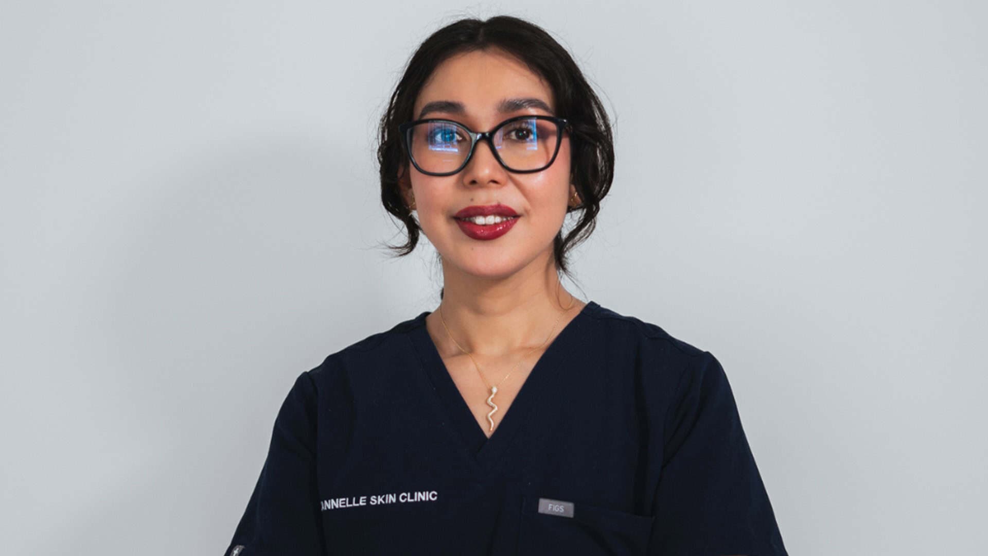 A woman with dark hair and glasses smiling in professional skincare uniform at Cannelle Skin Clinic.